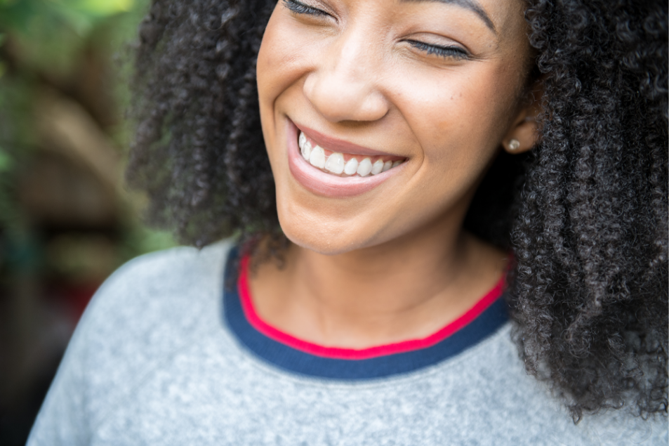 Face de uma mulher negra de cabelos cacheados com lindo sorriso de dentes brancos e árvores defocadas ao fundo.