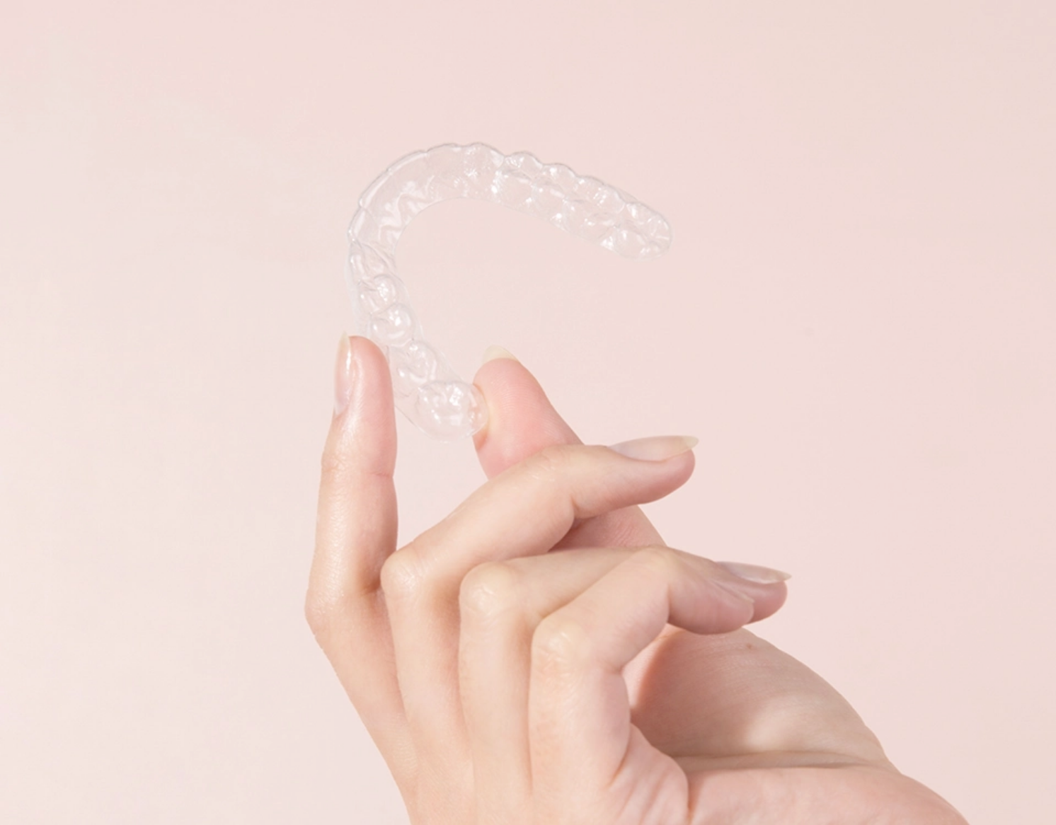 Mão segurando um aparelho dental transparente que quase se camufla em meio a frente de um fundo rosa bebê.