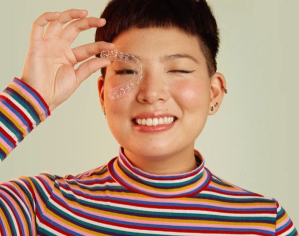 Mulher de aparência alternativa com um grande sorriso branco segurando o aparelho transparente em frente ao olho esquerdo.